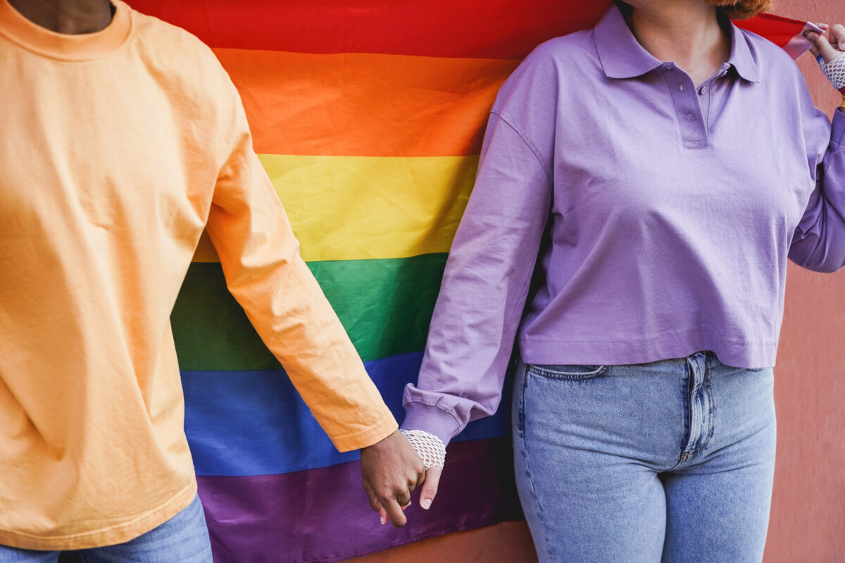 Happy women gay couple holding hands with rainbow flag on background