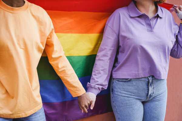 Happy women gay couple holding hands with rainbow flag on background